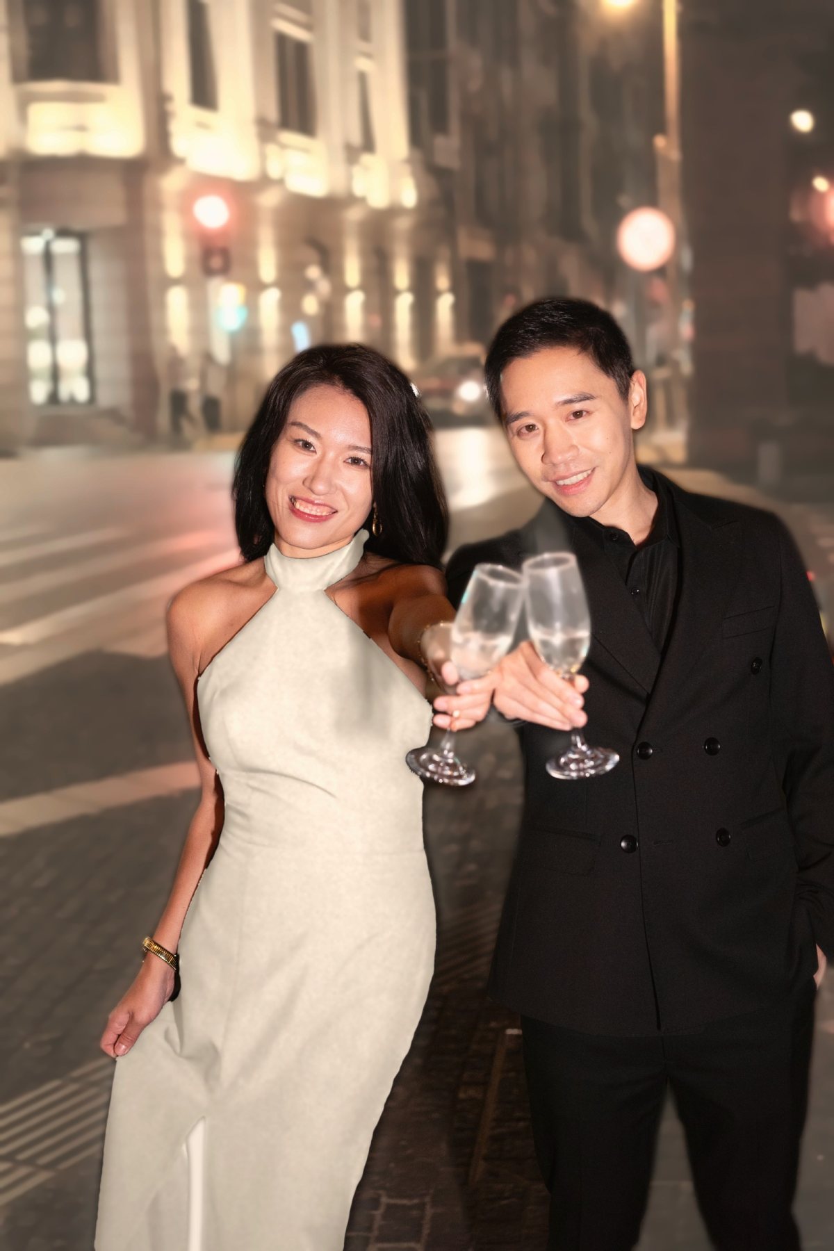Lan and Albert toasting with champagne on a softly lit city street, Lan in an ivory halter dress